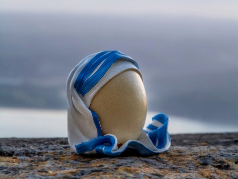 Œuf décoré d’un voile blanc et bleu, inspiré de l’habit emblématique de Mère Teresa, posé sur un rocher au coucher du soleil.