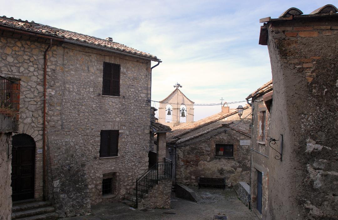 Glimpse of a village with stone houses and a view of a church’s bell-gable, amid narrow alleys and ancient rooftops.