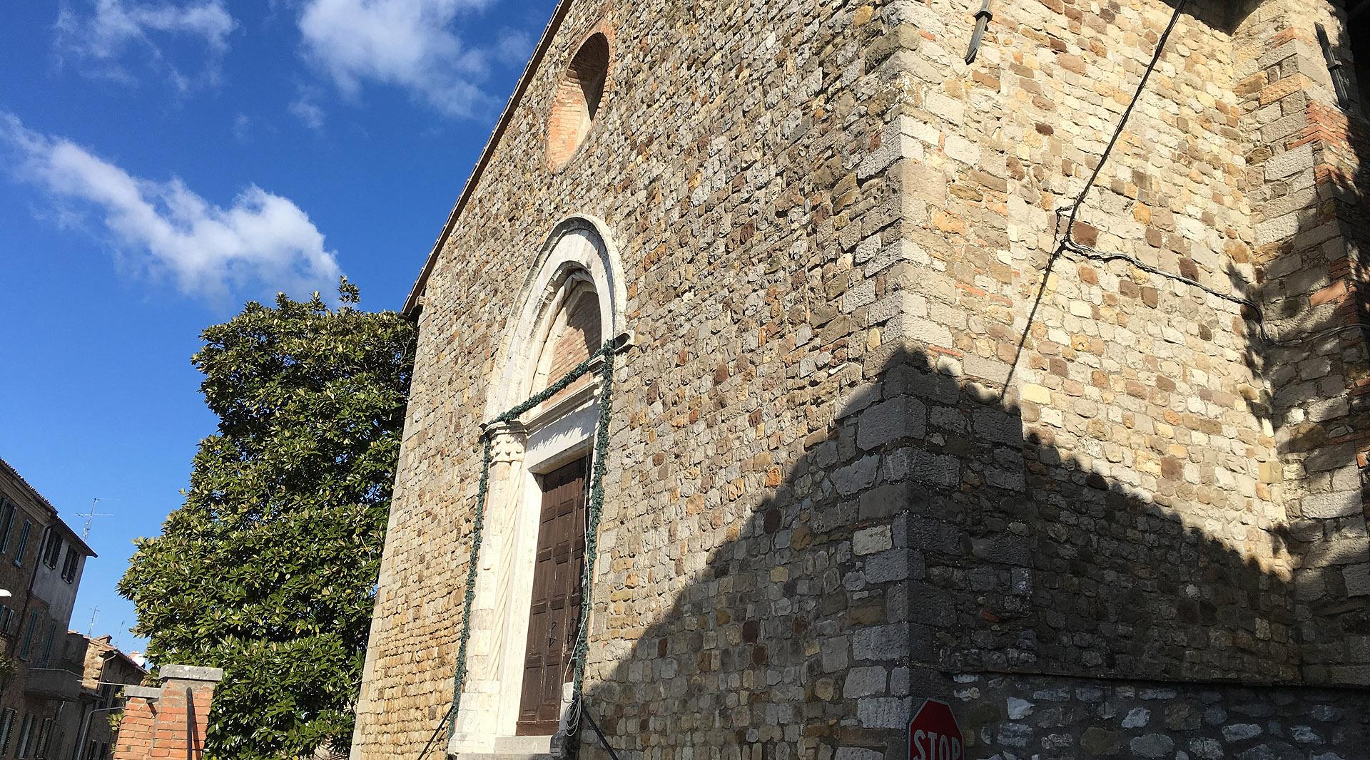 Façade en pierre de l’église Santa Maria Vecchia à Ficulle, éclairée par le soleil sous un ciel bleu.