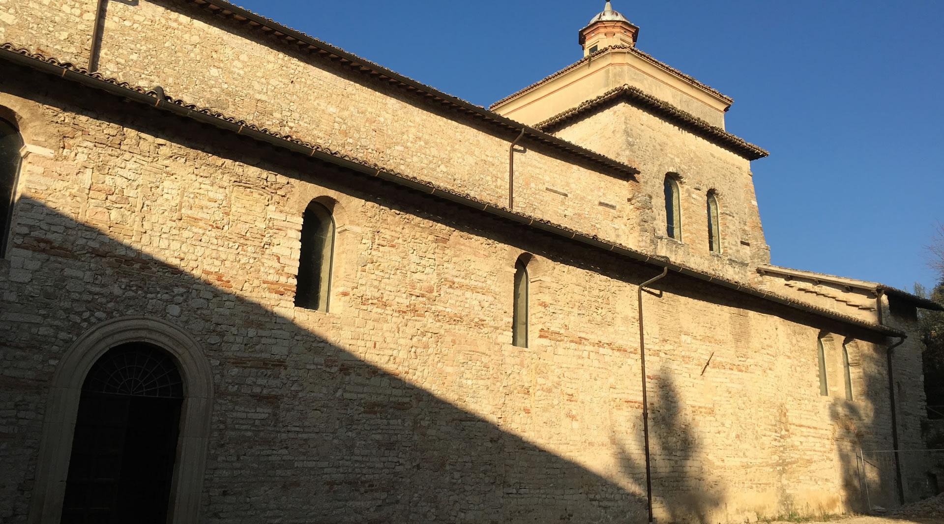 Exterior of the Basilica of San Salvatore in Spoleto, with stone walls, narrow windows, and an apse lit by sunlight.