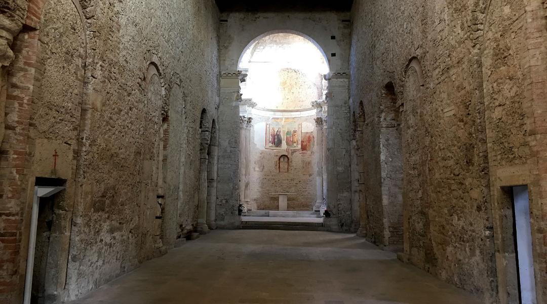Navata centrale in pietra della basilica di San Salvatore, con abside affrescata e luce naturale che illumina l’altare.