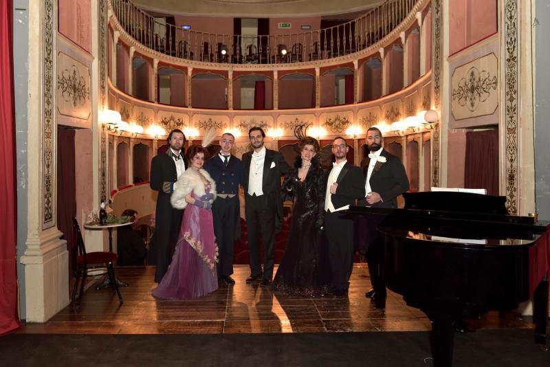 Vista dal palco del Teatro Cesare Caporali, che mostra la platea e il loggione in ferro. La sala interamente in legno è dipinta con una vernice chiara, arricchita da eleganti stucchi dorati. In primo piano artisti di scena.