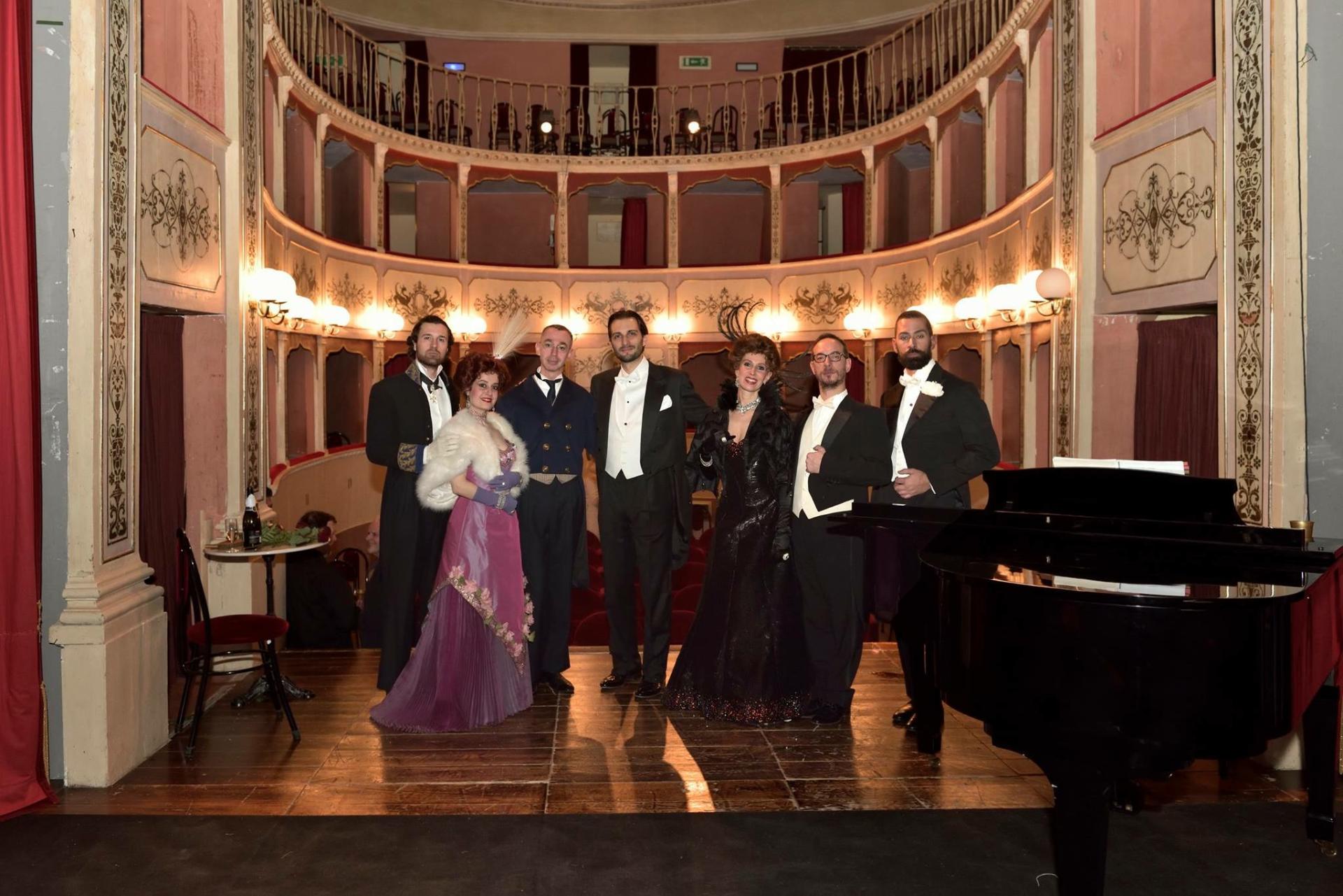 View from the stage of Teatro Cesare Caporali, showing the audience area and the iron balcony. The room, entirely made of wood, is painted with light-colored paint and enriched with elegant golden stucco. In the foreground, performers are on stage.