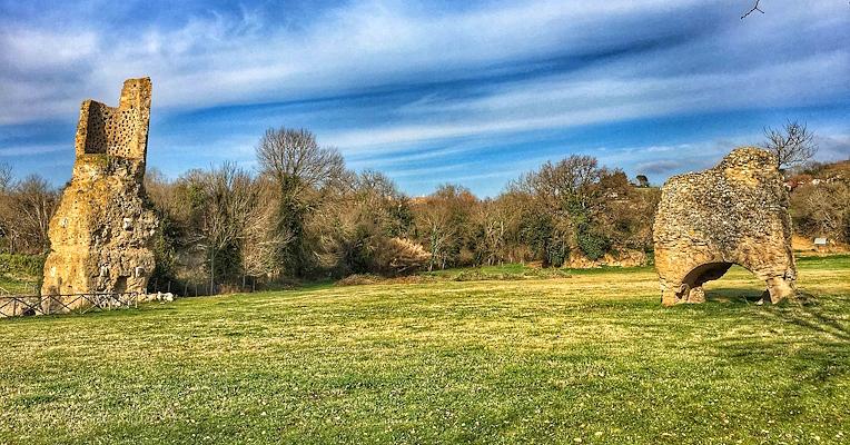 Remains of Roman structures on a green lawn, with trees in the background and a blue sky with scattered clouds