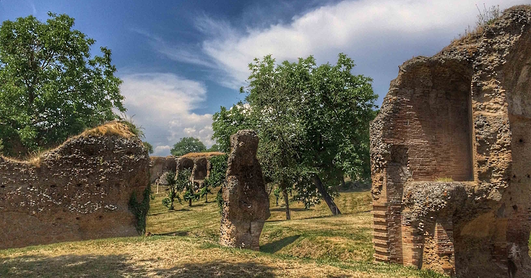 Vestiges de structures romaines sur une pelouse verte, avec des arbres à l’arrière-plan et un ciel bleu traversé de nuages