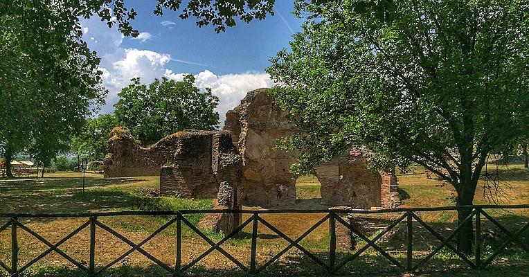 Remains of Roman structures among trees and vegetation, behind a wooden fence, illuminated by sunlight