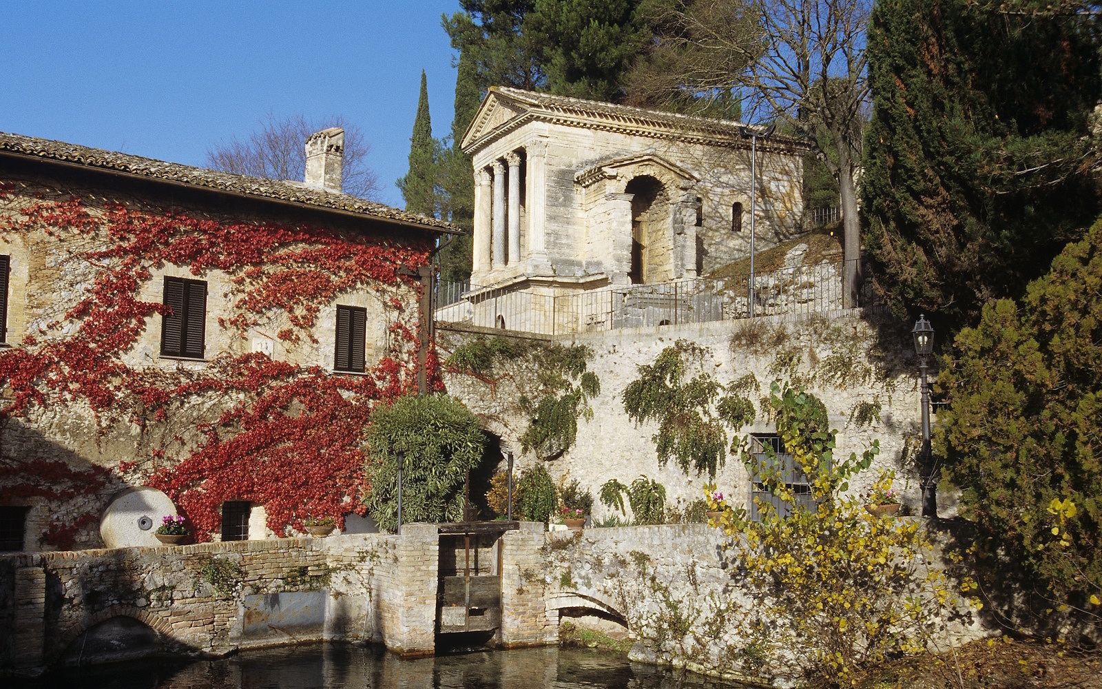 Il Tempietto sul Clitunno in alto e alle spalle di edificio coperto di edera rossa accanto a un canale