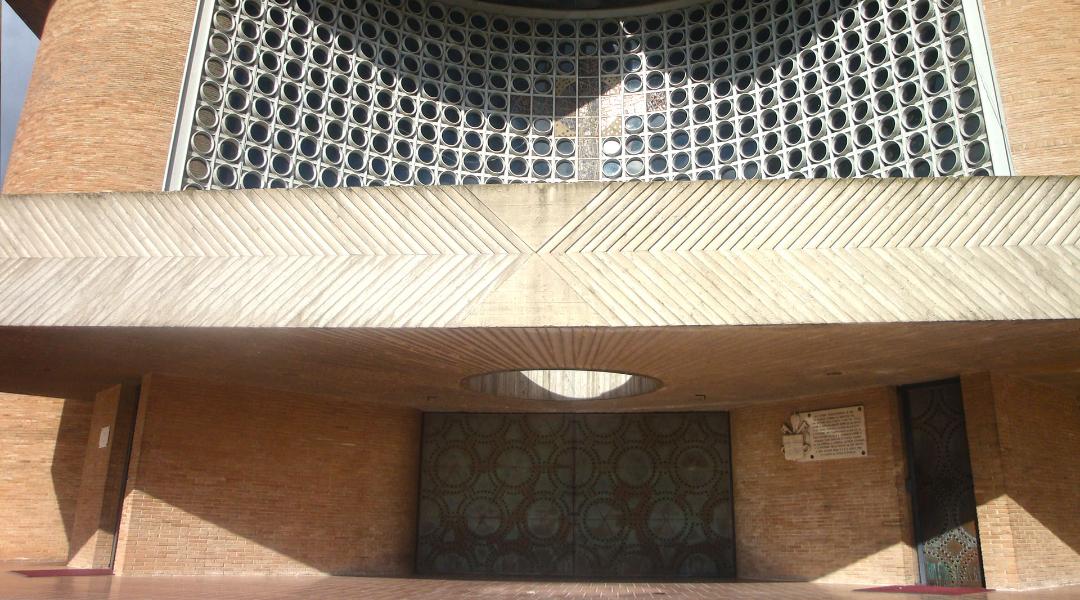 Bronze portal of the Collevalenza Sanctuary, with an overhanging perforated wall made of glass and concrete and geometric details.