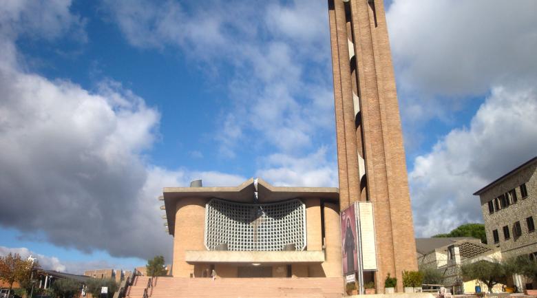  Modern facade of the Collevalenza Sanctuary with a tall brick bell tower, a blue sky, and clouds in the background. 
