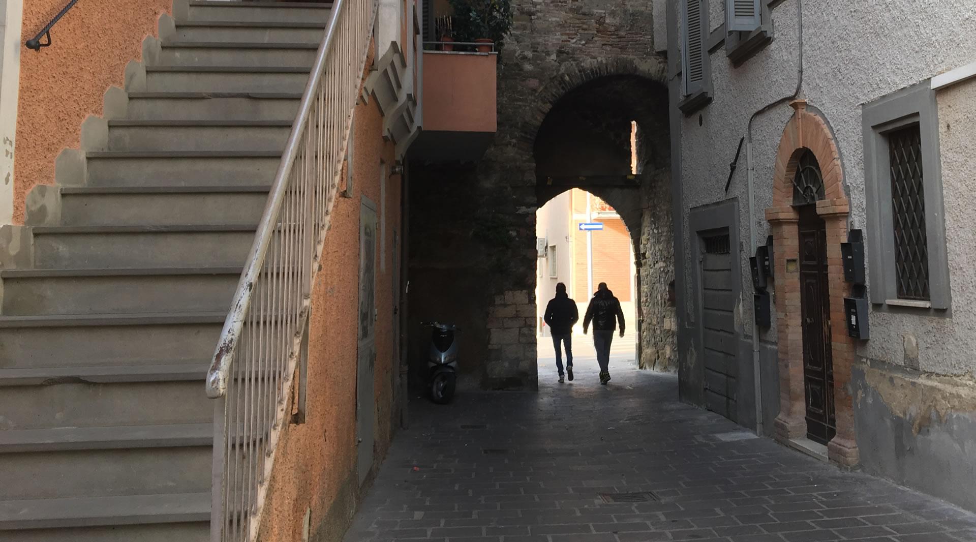 Deux personnes marchent sous l'arche en pierre de la Porta Sant'Angelo dans une ruelle étroite et pavée, entourée de bâtiments historiques aux façades en pierre et en plâtre. Un escalier extérieur en pierre est visible sur la gauche.