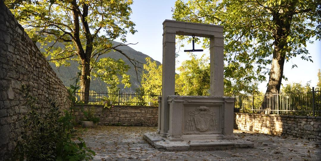 A historic stone well with an architrave and the noble coat of arms of the Poli family, surrounded by trees and walls in Usigni, Poggiodomo.