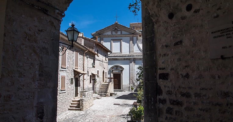 View of the Church of San Salvatore in Usigni through a stone arch, with historic houses and a cobbled street.