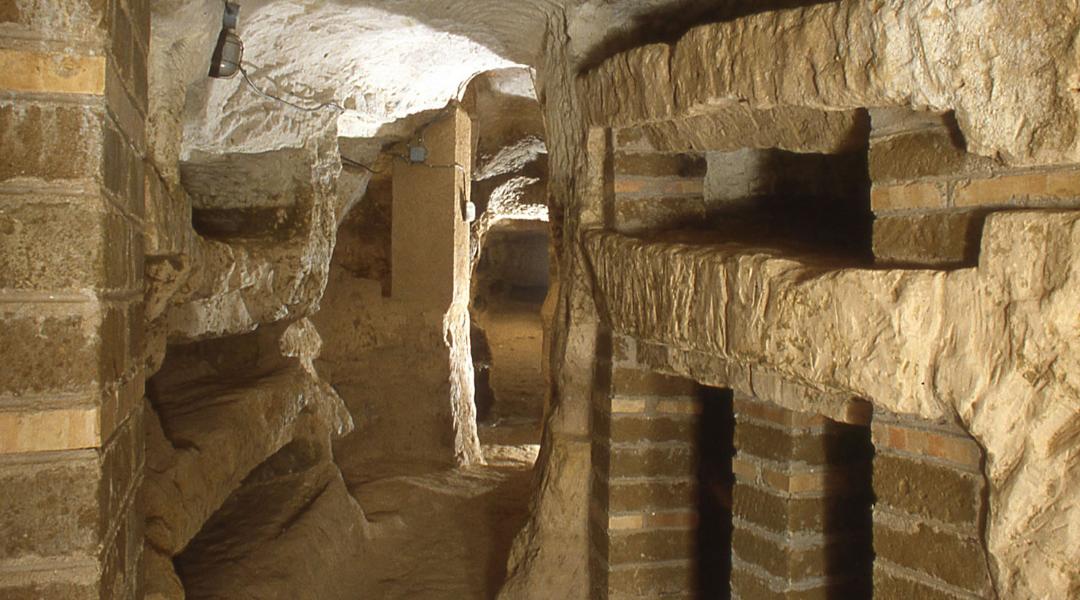 Interno della catacomba di Villa San Faustino. In primo piano si osserva un corridoio sotterraneo con pareti di roccia naturale, dove sono scavati diversi loculi di varie dimensioni. Il corridoio si apre verso una scala con un corrimano in ferro, che conduce a un livello superiore.