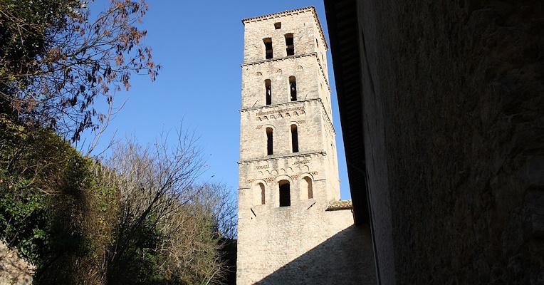 Bell tower of the Abbey of San Pietro in Valle in Ferentillo seen from below, with trees and blue sky in the background. The bell tower, with a quadrangular plan, is divided into five floors with arched windows and brick details.