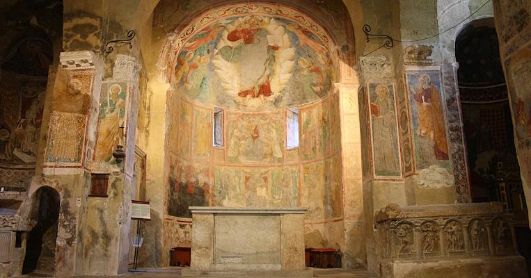 View of a richly decorated apse with frescoes depicting religious scenes and the main altar of the abbey, simple and central, with a marble structure.