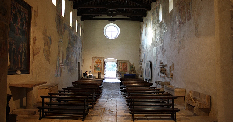 Interior of the abbey with a view towards the entrance and the circular rose window above the entrance. The central nave is flanked by wooden benches facing the altar, while the walls are decorated with frescoes.