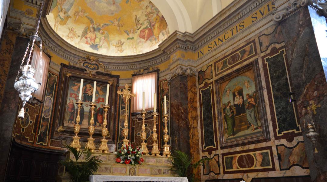 Primo piano dell'altare adornato con candelabri, un crocifisso dorato e un turibolo. Sullo sfondo dell'abside si ammira un’opera del Perugino: la 