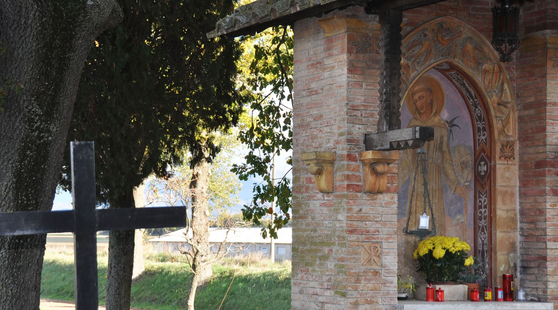 Votive aedicule with a painting of Saint Francis, yellow flowers and red candles, among trees and greenery.