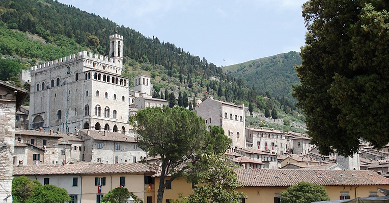 View of Gubbio, a medieval Umbrian village with the Palazzo dei Consoli, surrounded by stone houses and green hills.