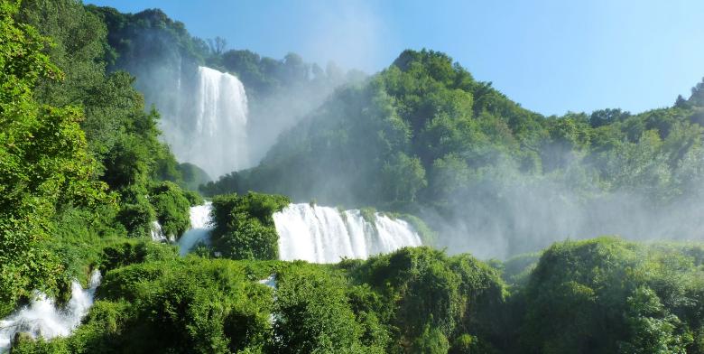 Immagine: The Marmore Waterfall seen from below: cascades of water among dense vegetation, with spray and mist rising towards the clear sky. 