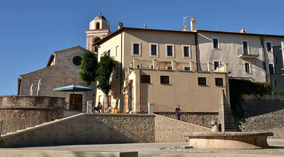 Square in Sellano with a fountain in the foreground, historic buildings, and the church with its bell tower in the background on a sunny day.