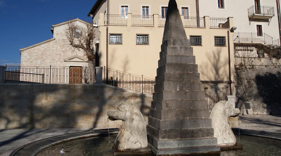 Fountain of Sellano with a central obelisk, with historic buildings and a stone church in the background.