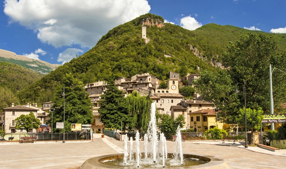 Square of Scheggino with a fountain, stone houses, and a medieval tower on a green hillside, under a blue sky with clouds.