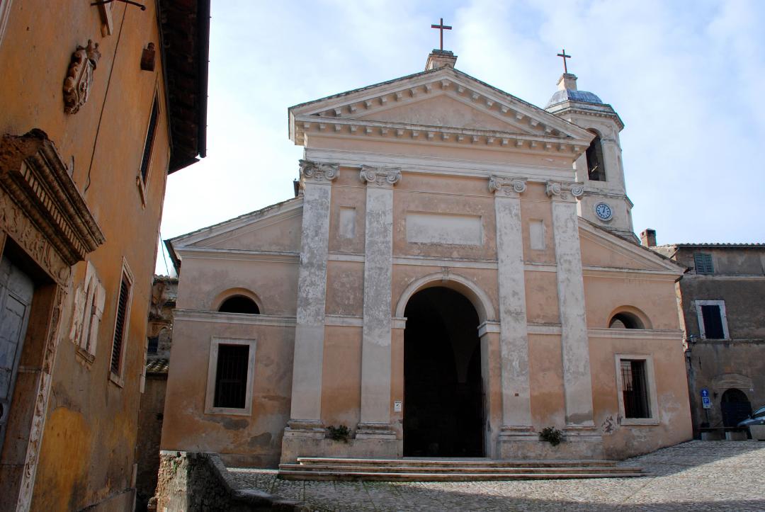 Façade of the Collegiate Church of Santa Maria Assunta in Otricoli, with bell tower and crosses, in a cobbled square.
