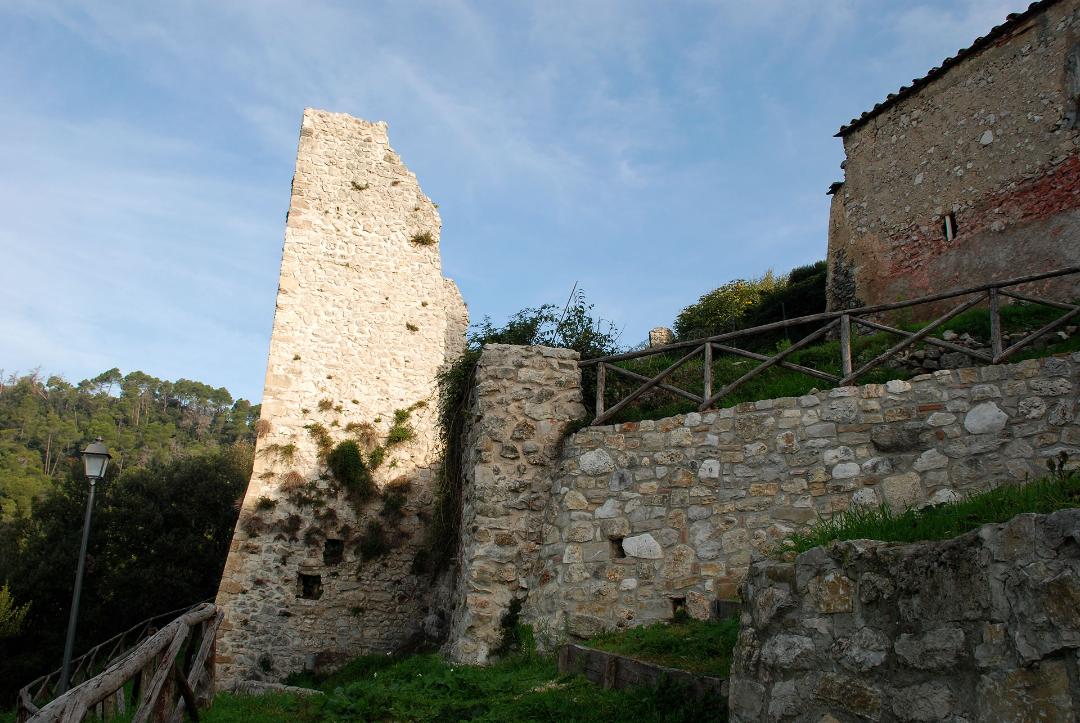 Remains of a medieval stone tower in Otricoli, with adjacent walls and a grassy path at the foot of the hill.