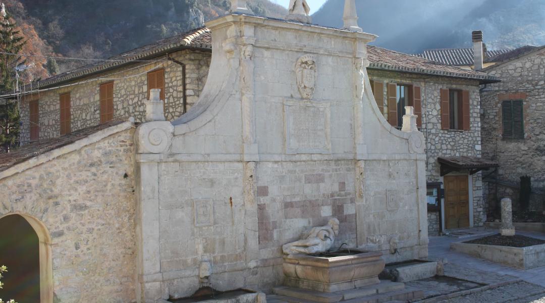 Stone fountain in Polino, adorned with sculptures and coats of arms; in the background, stone buildings with wooden shutters and mountains.