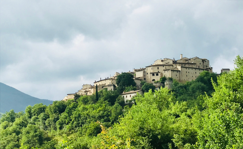 View of Vallo di Nera, a medieval village perched on a green hill with stone houses, under a cloudy sky.