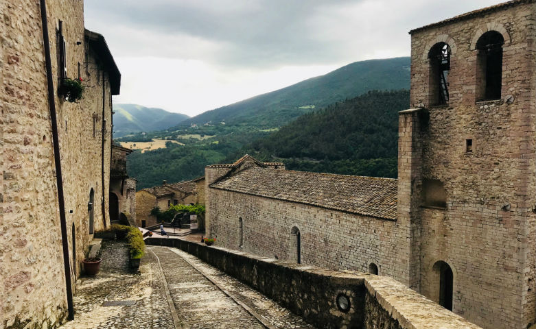 Stone-paved street among ancient buildings in a medieval village, with a bell tower and a panoramic view of hills and valleys.
