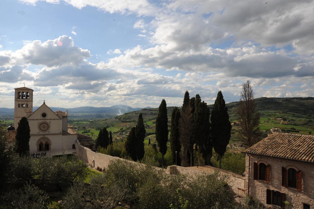 Panoramic view of Assisi with the Basilica of Saint Francis surrounded by cypress trees and green hills under a cloudy sky