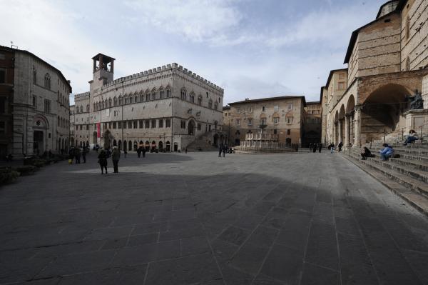  Historischer Platz in Perugia mit mittelalterlichen Gebäuden, einem monumentalen Brunnen in der Mitte und Menschen, die gehen oder auf den Stufen sitzen. 