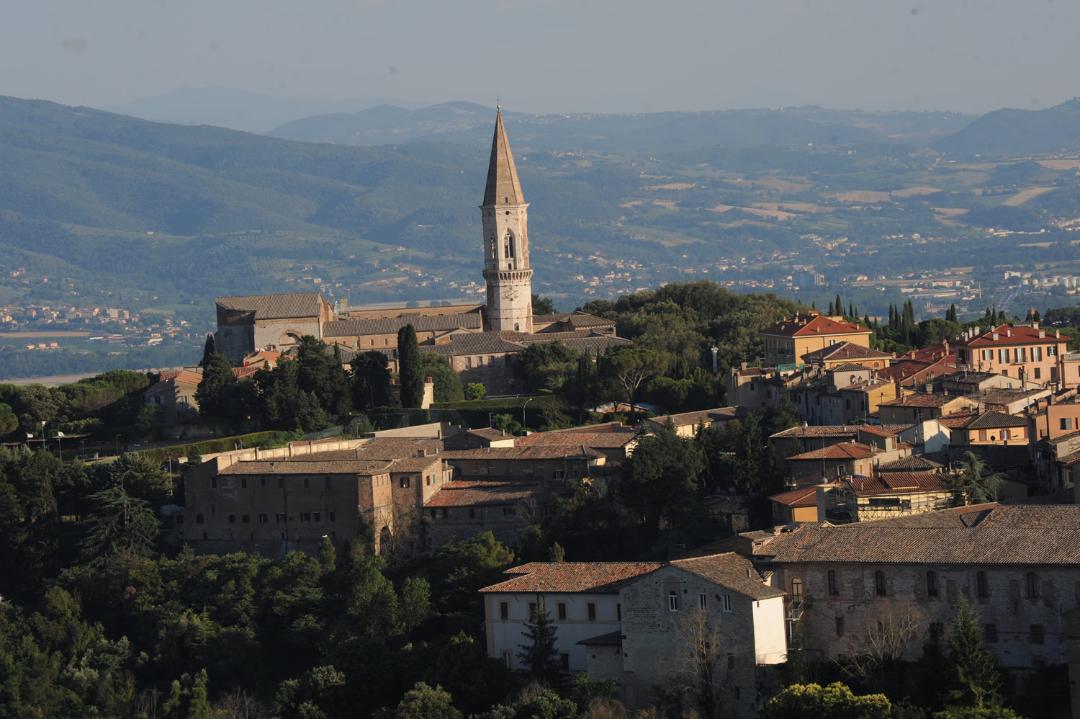 Hilltop view with the cathedral and its tall bell tower rising among historic buildings, with the Umbrian mountains in the background.