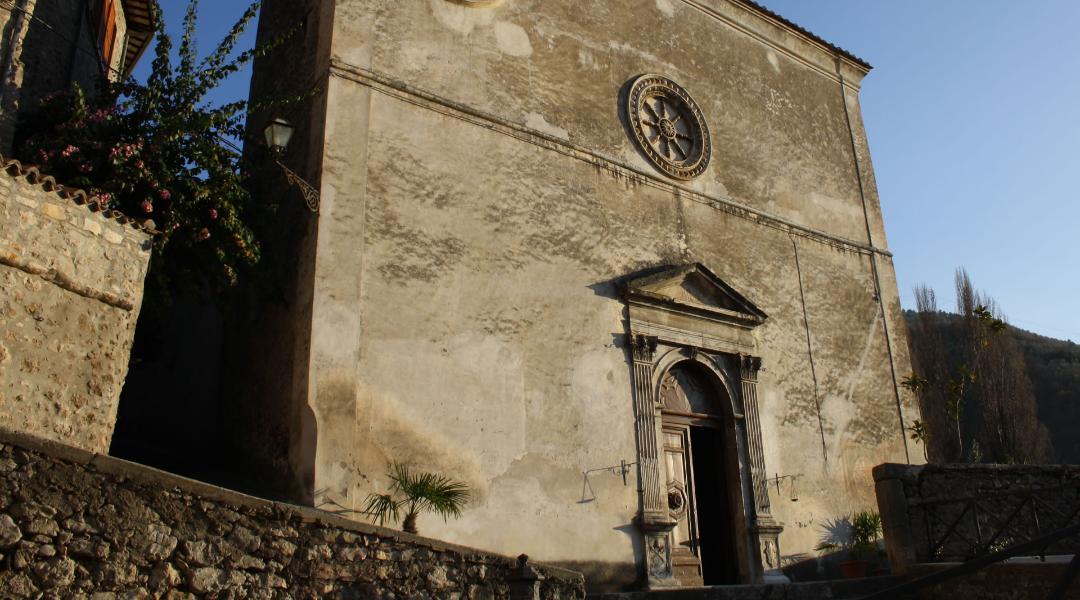 Ancient church with a weathered facade, stone portal, and rose window, set among stone walls and lush vegetation.
