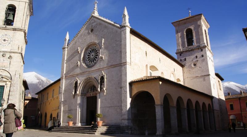Basilica di San Benedetto a Norcia, con la sua facciata gotica e il rosone, incorniciata da montagne innevate sotto un cielo limpido.