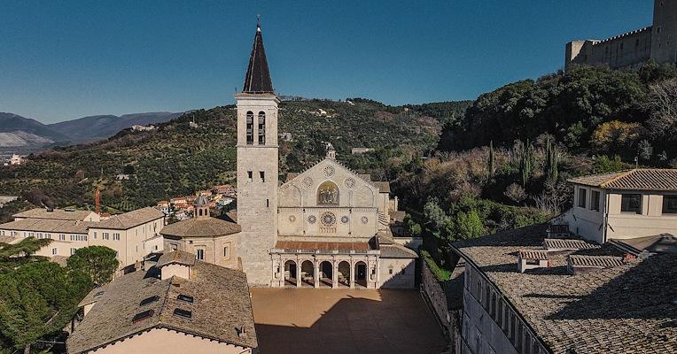 Veduta aerea del Duomo di Spoleto con facciata romanica, campanile e piazza, circondato da colline e tetti storici.
