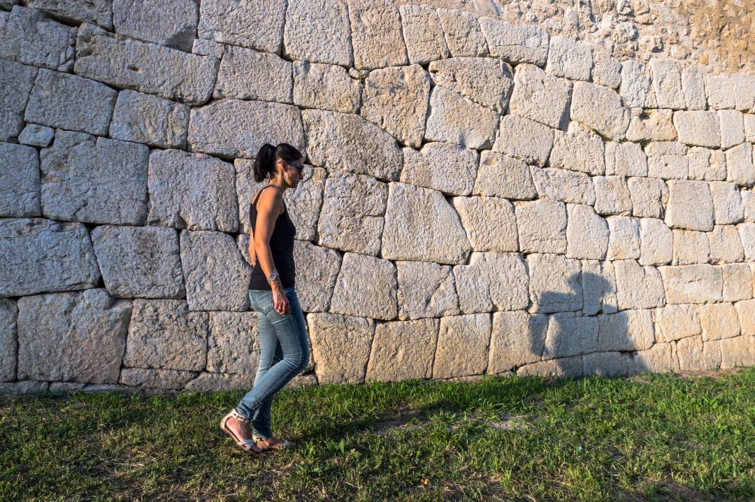 A section of the opus polygonal defensive wall of Amelia with a woman walking close to the wall and casting her shadow on the large irregularly squared blocks that make up the wall face.