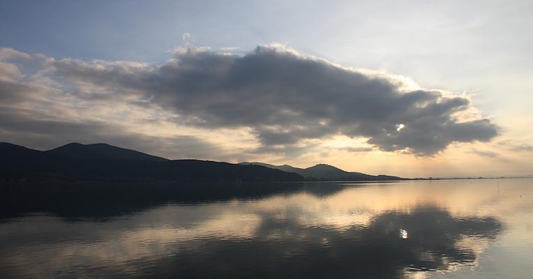 A scenic view of Lake Trasimeno at sunset, with clouds reflecting on the calm water and mountains in the background.