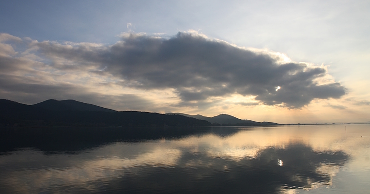 Ein malerischer Blick auf den Trasimenischen See bei Sonnenuntergang, mit Wolken, die sich im ruhigen Wasser spiegeln, und Bergen im Hintergrund.