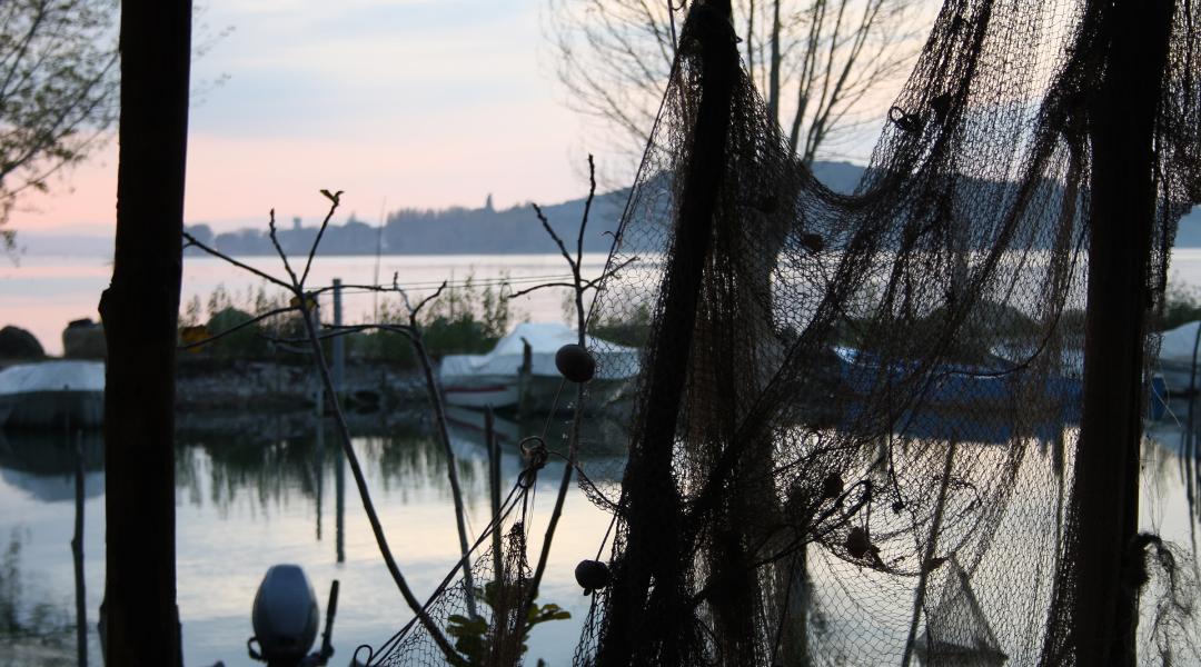 Hanging fishing nets overlooking Lake Trasimeno at sunset, with moored boats and the silhouette of a village in the distance.