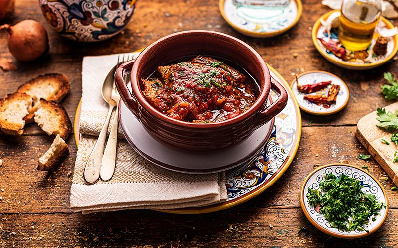 Umbrian tegamaccio served in terracotta, lake fish stewed in tomato sauce with parsley, rustic bread, and spices on a decorated wooden table.