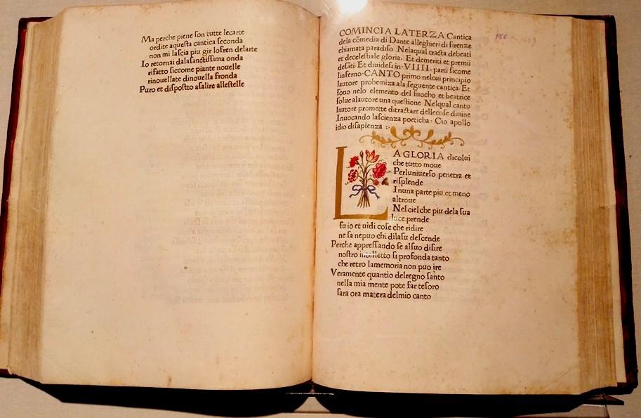Ancient manuscript of the Divine Comedy opened, featuring a decorated initial and golden ornamental details.