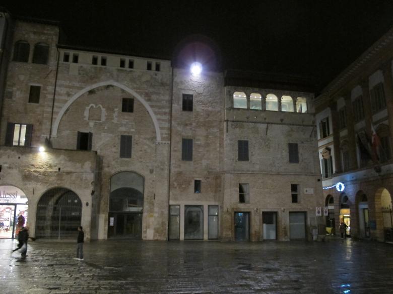 Immagine: Historic brick building with a pointed arch and an illuminated loggia, at night, in a square. 