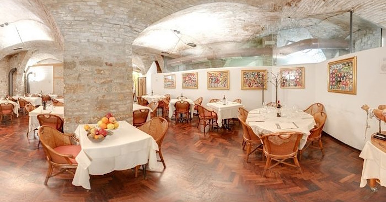 Restaurant hall with a vaulted stone ceiling, paintings on the walls, wooden flooring, and set tables.