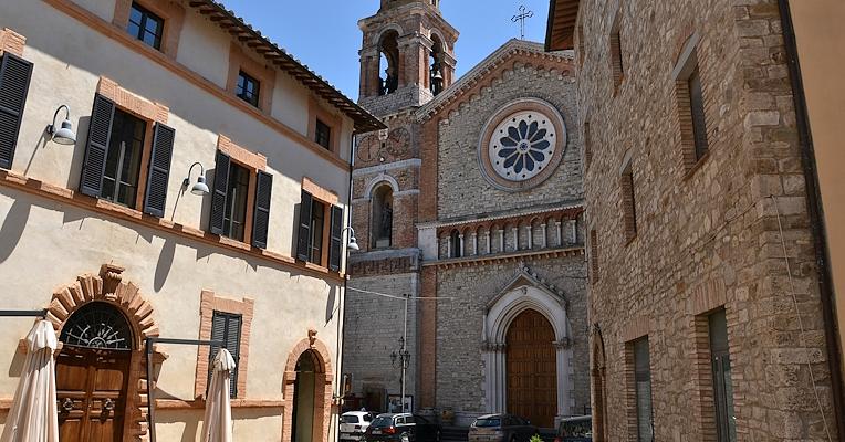 Vista di una piazza con edifici in pietra e mattoni, finestre con persiane scure e una chiesa con rosone e campanile sullo sfondo.