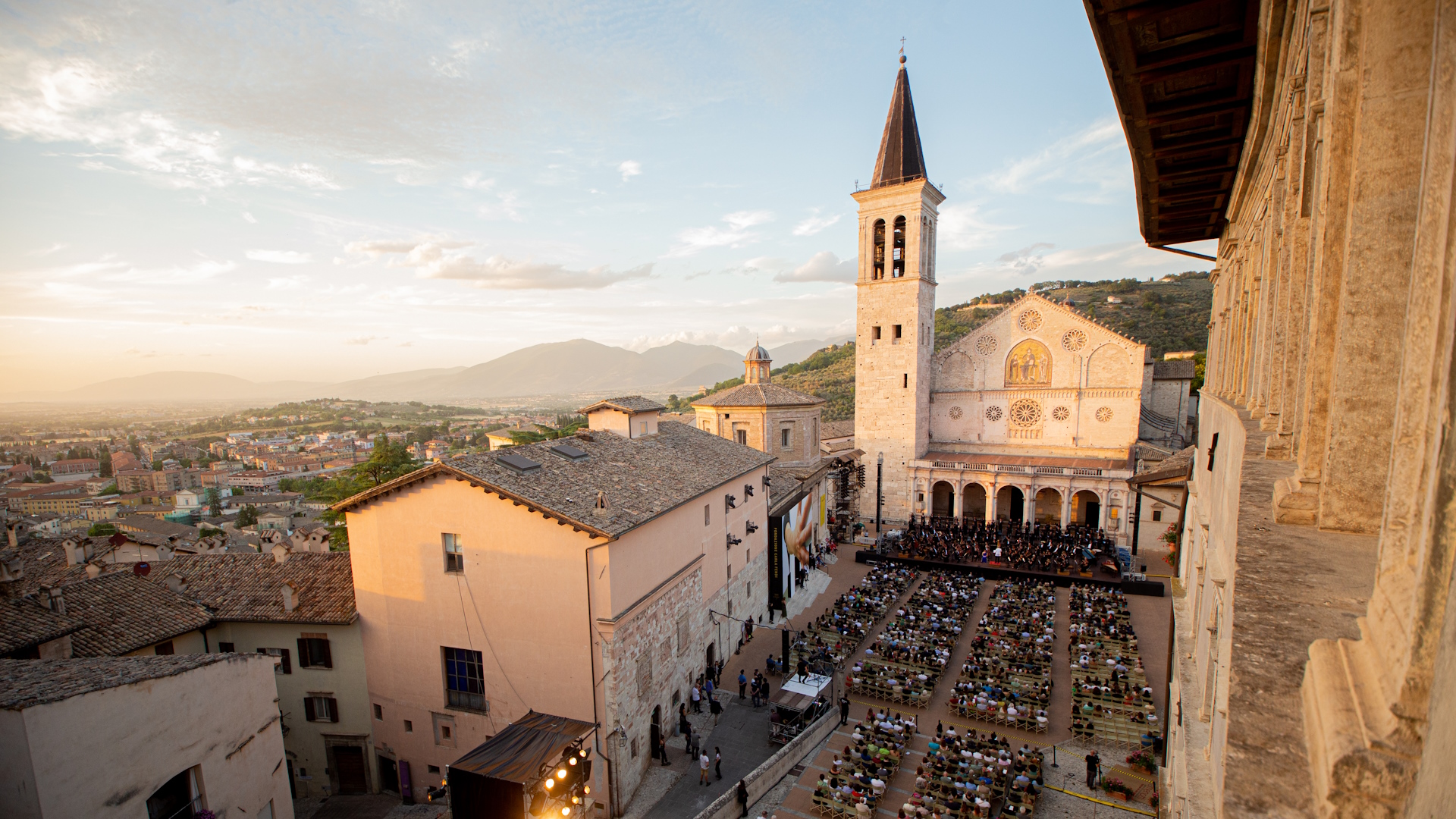 View of Piazza Duomo in Spoleto during a concert of the Festival dei Due Mondi, with the audience seated in front of the illuminated Cathedral at sunset.
