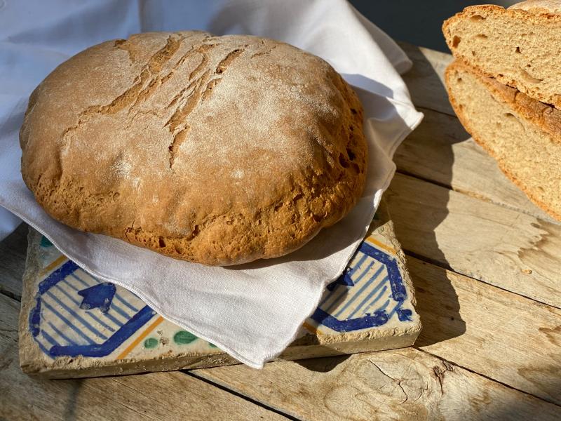 Pagnottella rustica di farro su mattonella decorata, affiancata da fette di pane, illuminata da luce naturale.