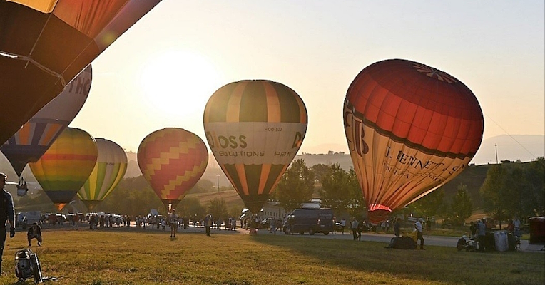 Un groupe de personnes s’affaire à faire décoller des montgolfières posées sur une prairie, alors que le soleil se lève dans le ciel